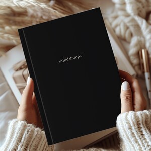 Puede incluir: Un diario negro con las palabras "mind dumps" en una fuente minimalista. El diario está abierto, revelando las páginas en el interior. La imagen está colocada sobre un fondo suave y neutro, con un bolígrafo y otros objetos visibles.
