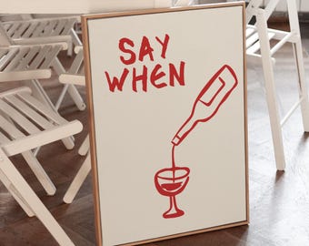 May include: A framed canvas print with the words "SAY WHEN" in red, along with a red illustration of a wine bottle pouring into a wine glass. The artwork has a light beige background and a wooden frame. The image is set in a dining room.
