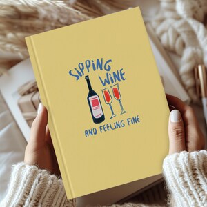 May include: A yellow notebook with the text "Sipping Wine and Feeling Fine" in blue and black lettering. The notebook features a hand-drawn illustration of a bottle of wine and two wine glasses.