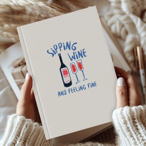 May include: A white notebook with the text "Sipping Wine and Feeling Fine" in blue lettering. The notebook features a hand-drawn illustration of a bottle of wine and three wine glasses.