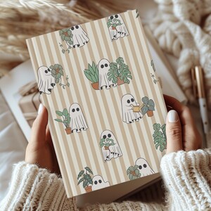 May include: A beige and white striped journal featuring a repeating pattern of cartoon ghosts with plants. The ghosts are white with black eyes, and the plants are green and brown. The journal is held open, revealing blank pages.