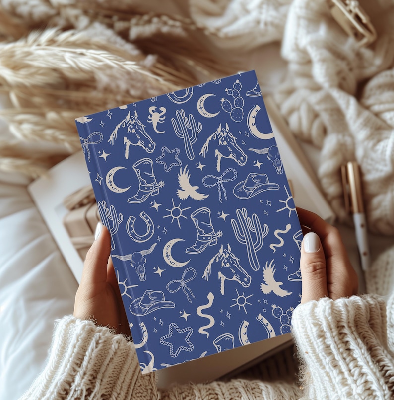 Puede incluir: Una libreta azul con un patr&oacute;n blanco de ilustraciones de tem&aacute;tica western. Las ilustraciones incluyen caballos, cactus, estrellas, botas y herraduras.