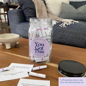 May include: A clear glass jar labeled "You Got This" filled with rolled-up affirmations. The jar has a black lid and sits on a wooden surface. Affirmations are scattered around the jar. The text on the jar reads "100 affirmations to uplift, encourage, and inspire every day."
