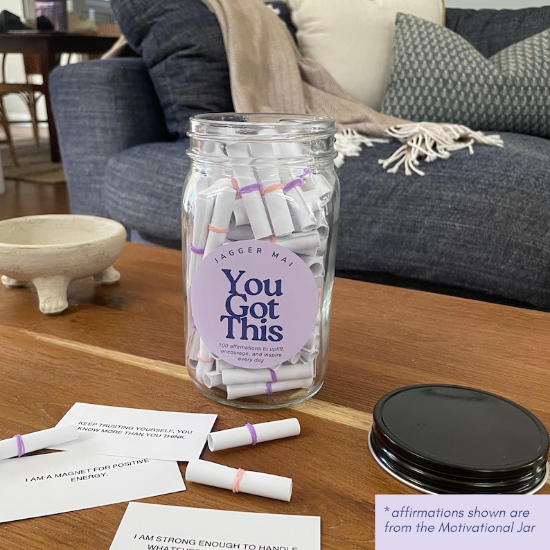 May include: A clear glass jar labeled "You Got This" filled with rolled-up affirmations. The jar has a black lid and sits on a wooden surface. Affirmations are scattered around the jar. The text on the jar reads "100 affirmations to uplift, encourage, and inspire every day."