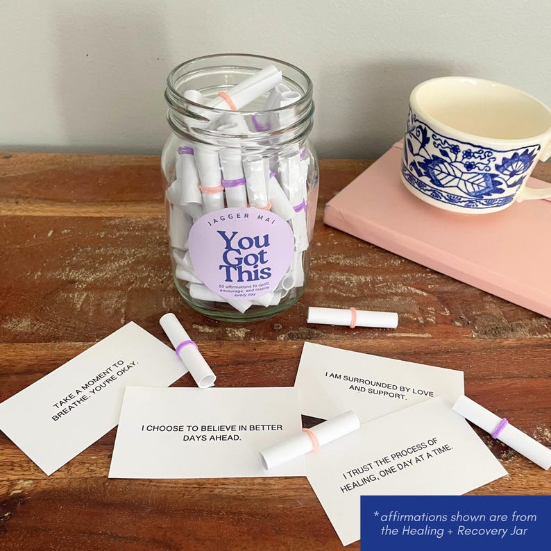May include: A clear glass jar filled with rolled-up white paper scrolls, secured with pink and purple rubber bands. The jar has a purple label that reads "You Got This." Several affirmation cards are scattered around the jar, with phrases like "Take a moment to breathe, you're okay."
