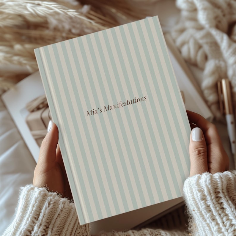 May include: A journal with a striped pattern in light blue and beige, titled "Mia's Manifestations." The journal is held open, revealing a glimpse of the inside pages. The cover design features vertical stripes.