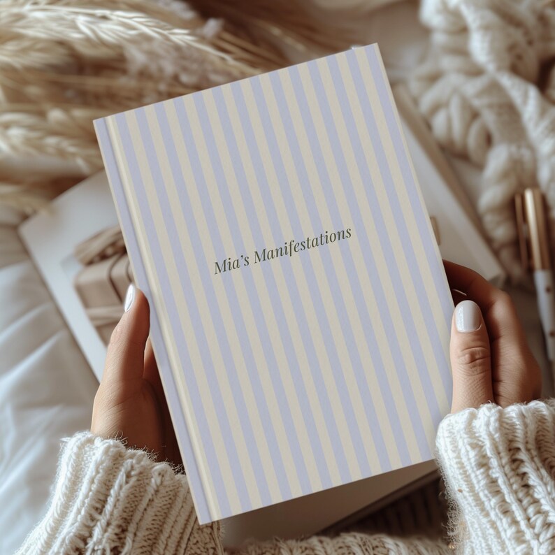 May include: A journal with a light blue and cream vertical striped cover. The text "Mia's Manifestations" is printed in a dark gray font. The journal is held open in a person's hands, with a soft, neutral-toned background.