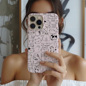 May include: A pink phone case featuring a repeating pattern of various cat illustrations in black. The cats are depicted in different poses and expressions, creating a playful and whimsical design. The case is shown held in front of a person.