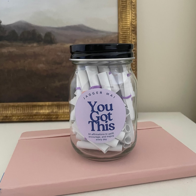 May include: A clear glass jar with a black lid, filled with rolled-up white papers secured with purple bands. A round label on the jar reads "You Got This" with the text "50 affirmations to uplift, encourage, and inspire every day."