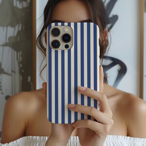 May include: A smartphone case with a vertical striped pattern in navy blue and cream. The case has a camera cutout and is held in front of a person's face.
