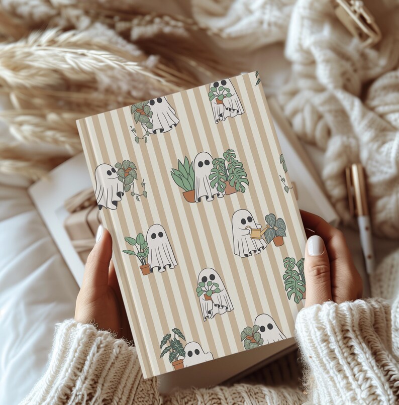 May include: A beige and white striped journal featuring a repeating pattern of cartoon ghosts with plants. The ghosts are white with black eyes, and the plants are green and brown. The journal is held open, revealing blank pages.