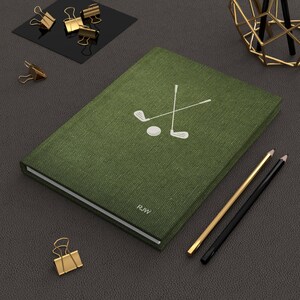 K&ouml;nnte beinhalten: Ein gr&uuml;nes, mit Leinen bezogenes Notizbuch mit einem wei&szlig;en Golfclub- und Ball-Design. Das Notizbuch tr&auml;gt die Initialen "R.W." auf dem Einband.