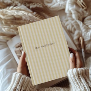 May include: A hardcover journal with a cream and yellow striped cover. The text "Mia's Manifestations" is printed on the cover. The journal is open, revealing blank pages. A pen rests nearby. The journal is held by a person wearing a cream-colored sweater.