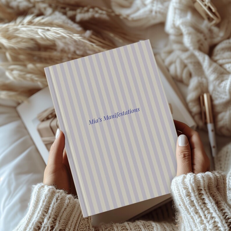 May include: A journal with a striped cover in light purple and cream colors. The text "Mia's Manifestations" is printed in a dark blue script. The journal is held open, revealing blank pages.