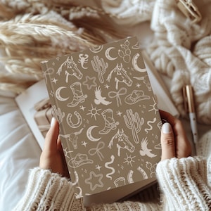 Puede incluir: Una libreta marr&oacute;n con un patr&oacute;n blanco de tem&aacute;tica western. El patr&oacute;n incluye cactus, herraduras, estrellas, lunas y botas de vaquero.
