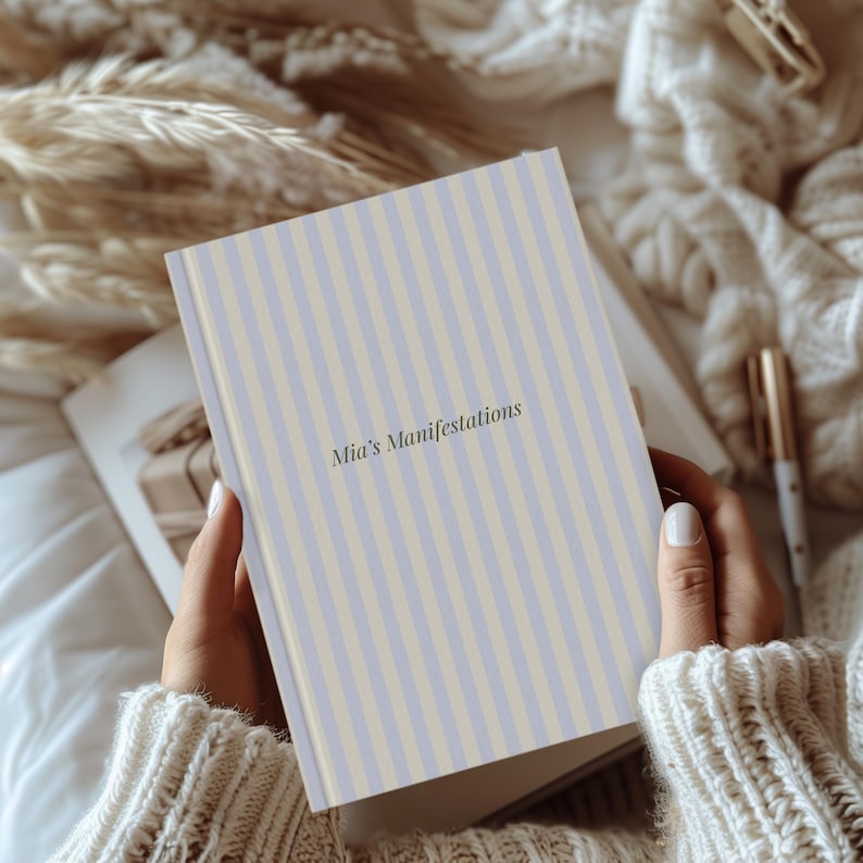May include: A journal with a light blue and beige striped cover. The text "Mia's Manifestations" is printed in a soft green font. The journal is held open, revealing blank pages, and is surrounded by soft, neutral-toned decor.