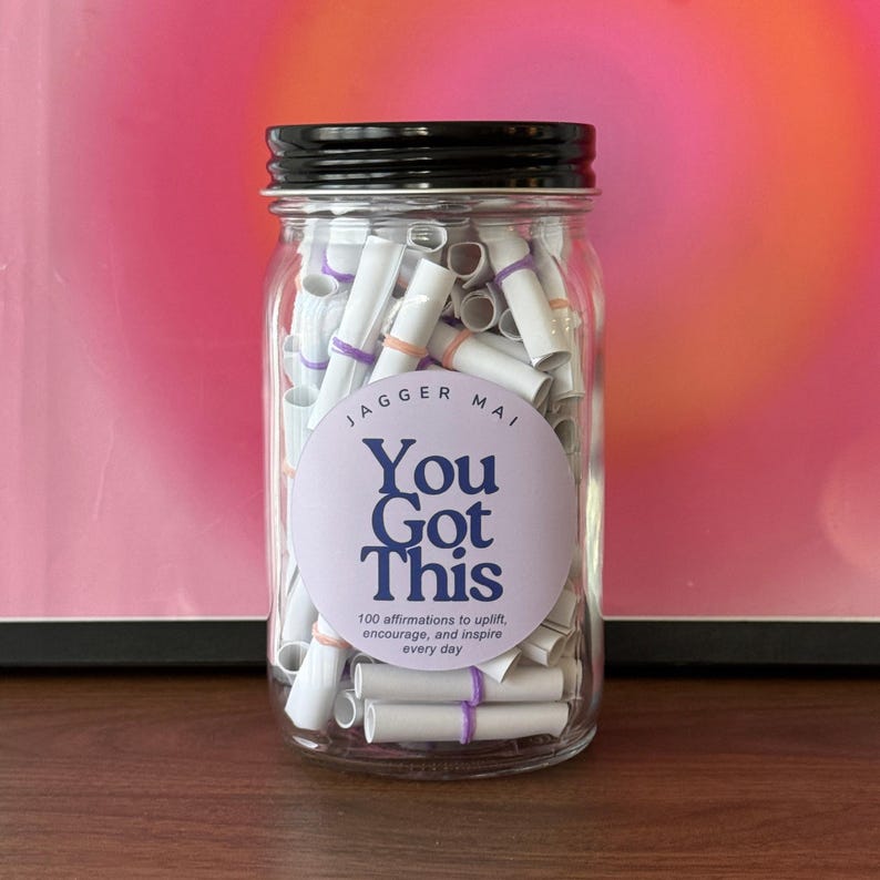 May include: A clear glass jar with a black lid filled with rolled-up paper scrolls. The jar has a round label with the text "You Got This" and "100 affirmations to uplift, encourage, and inspire every day". The scrolls are secured with purple and orange bands.