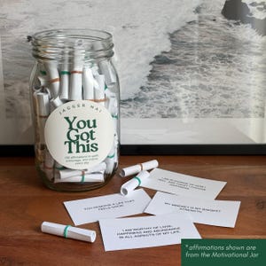 May include: A clear glass jar filled with rolled-up white paper slips, secured with green and brown rubber bands. The jar has a circular label that reads "You Got This". Several affirmation cards are scattered around the jar.