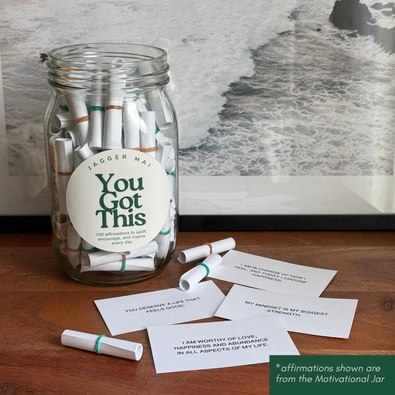 May include: A clear glass jar filled with rolled-up white paper slips, secured with green and brown rubber bands. The jar has a circular label that reads "You Got This". Several affirmation cards are scattered around the jar.