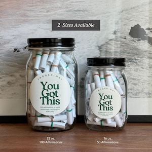 May include: Two glass jars with black lids filled with rolled-up white paper scrolls. The jars have labels that read "You Got This" and include the text "100 affirmations to uplift, encourage, and inspire every day." The larger jar is 32 oz and the smaller is 16 oz.