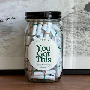 May include: A clear glass jar with a black lid filled with rolled-up white paper scrolls. A white circular label with the text "You Got This" in green is affixed to the jar. The label also includes the text "100 affirmations to uplift, encourage, and inspire every day."