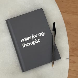 May include: A grey notebook with the words "notes for my therapist" in white. A black pen lies beside the notebook. The notebook and pen are on a white marble surface.