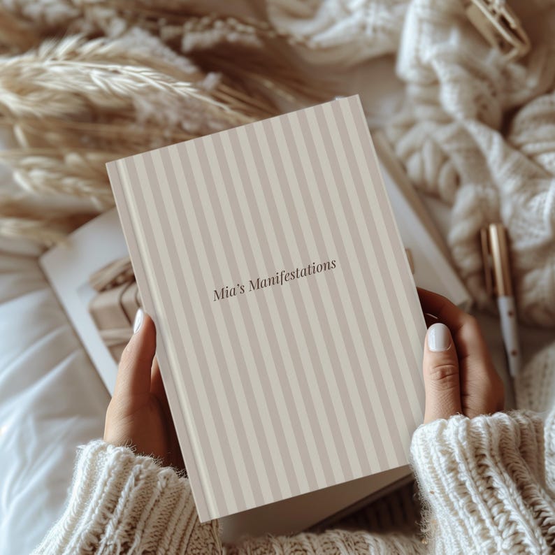 May include: A journal with a beige and white striped cover, titled "Mia's Manifestations." The book is held open, revealing pages inside. The image has a soft, neutral color palette, with a cozy, inviting aesthetic.