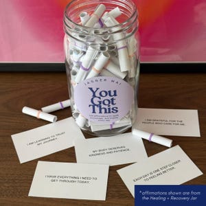May include: A clear glass jar filled with rolled-up white paper affirmations, secured with colorful bands. The jar has a label that reads "You Got This." Several affirmation cards are scattered around the jar, with phrases like "I am learning to trust my journey."