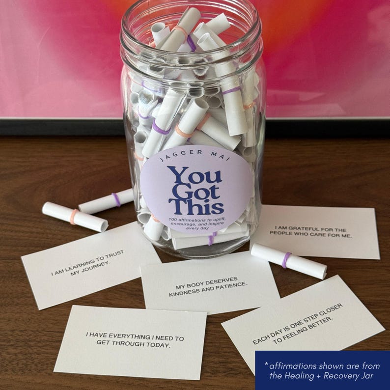 May include: A clear glass jar filled with rolled-up white paper affirmations, secured with colorful bands. The jar has a label that reads "You Got This." Several affirmation cards are scattered around the jar, with phrases like "I am learning to trust my journey."