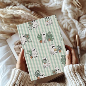 May include: A notebook with a light green and white striped background. The notebook features a repeating pattern of friendly ghost characters tending to potted plants. The cover is held open, revealing blank pages.