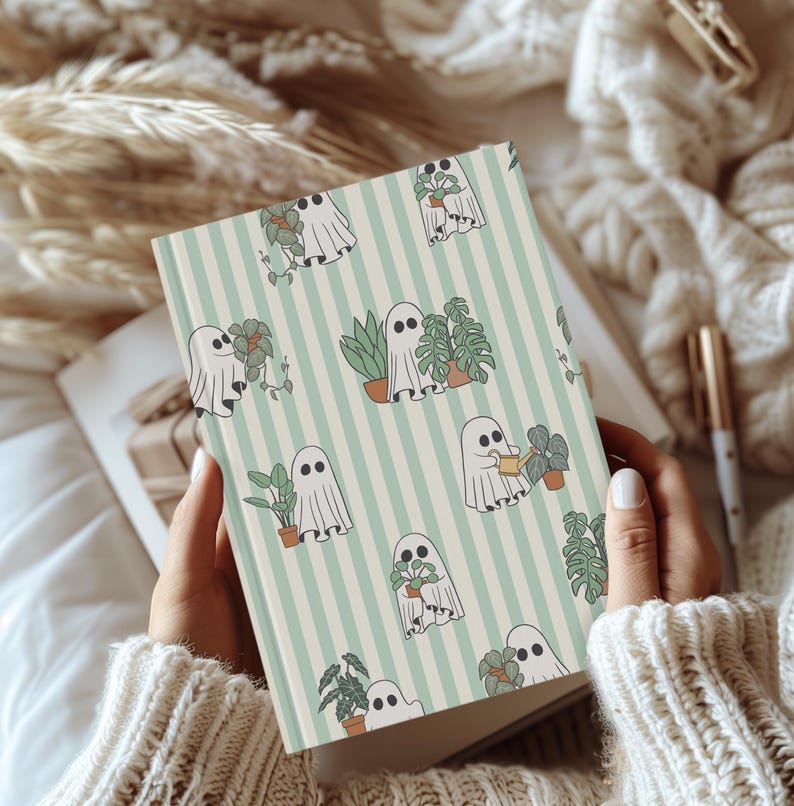 May include: A notebook with a light green and white striped background. The notebook features a repeating pattern of friendly ghost characters tending to potted plants. The cover is held open, revealing blank pages.