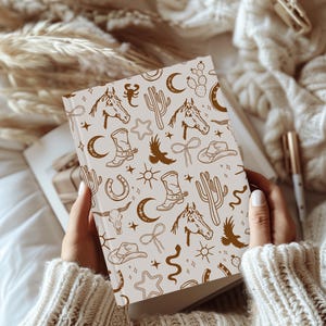 Puede incluir: Un cuaderno blanco con un patr&oacute;n marr&oacute;n y blanco de ilustraciones de tem&aacute;tica western. Las ilustraciones incluyen caballos, cactus, estrellas, botas y otros s&iacute;mbolos del oeste.
