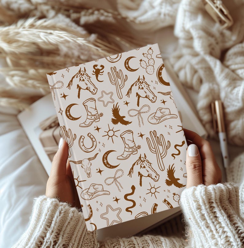 Puede incluir: Un cuaderno blanco con un patr&oacute;n marr&oacute;n y blanco de ilustraciones de tem&aacute;tica western. Las ilustraciones incluyen caballos, cactus, estrellas, botas y otros s&iacute;mbolos del oeste.