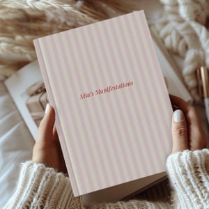 May include: A hardcover journal with a pink and cream striped cover. The text "Mia's Manifestations" is printed in a reddish-brown color. The journal is held in a person's hands, with a soft, textured sweater in the background.