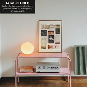 K&ouml;nnte beinhalten: Ein rosa Metallregal mit zwei Ablagen, einem wei&szlig;en Lautsprecher, einem Stapel B&uuml;cher und einer wei&szlig;en Globuslampe. An der Wand hinter dem Regal h&auml;ngt ein gerahmter Druck einer Collage aus Streichholzschachteln mit verschiedenen Texten und Bildern. Der Text auf den Streichholzschachteln lautet "Howdy", "This must be the place!" und "Hell Yeah".