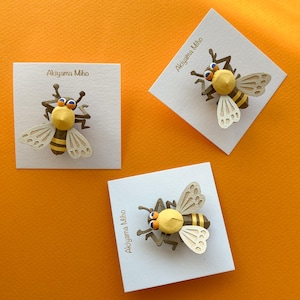 May include: Three handmade bee brooches on white cards against an orange backdrop. Each bee features a yellow head, a striped body, and detailed wings. The cards are labelled "Akiyama Miho".