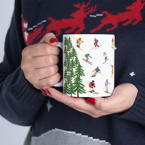 May include: White ceramic mug featuring a repeating pattern of skiers in colorful outfits and green pine trees. The mug is held by a person wearing a navy blue sweater with a red reindeer design.