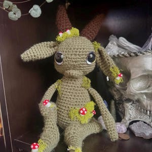 May include: A crocheted green goat with brown horns and red and white mushroom accents. The goat is sitting on a dark surface with a skull and crystals in the background.