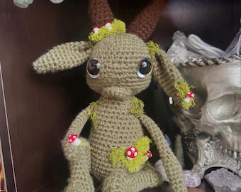 May include: A crocheted green goat with brown horns and red and white mushroom accents. The goat is sitting on a dark surface with a skull and crystals in the background.