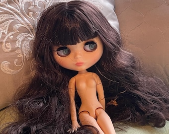 May include: A doll with a large head, pale skin, and long, wavy dark brown hair with bangs. The doll features large, forward-facing eyes with dark lashes, a small nose, and pink lips. It has an articulated body with visible joints at the shoulders and knees.