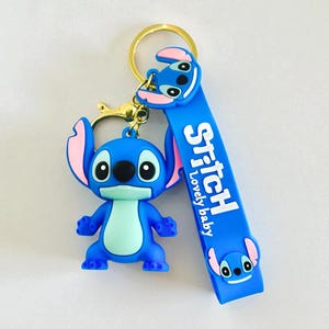 May include: A blue Stitch keychain with a gold-colored keyring. The keychain features a 3D Stitch figure and a blue strap with the word "Stitch" and "Lovely baby" printed on it. The ears are pink.