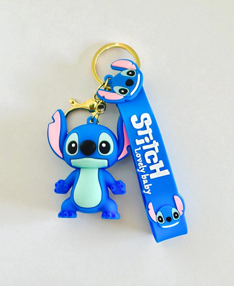 May include: A blue Stitch keychain with a gold-colored keyring. The keychain features a 3D Stitch figure and a blue strap with the word "Stitch" and "Lovely baby" printed on it. The ears are pink.
