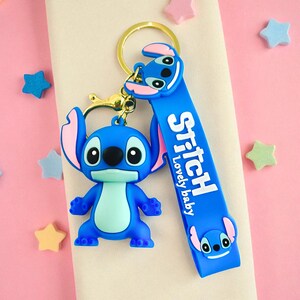 May include: A blue Stitch keychain with a gold-colored keyring and clasp. The keychain features a 3D Stitch figure and a blue strap with the word "Stitch" and "Lovely baby" printed on it. Pink and blue stars are scattered around.
