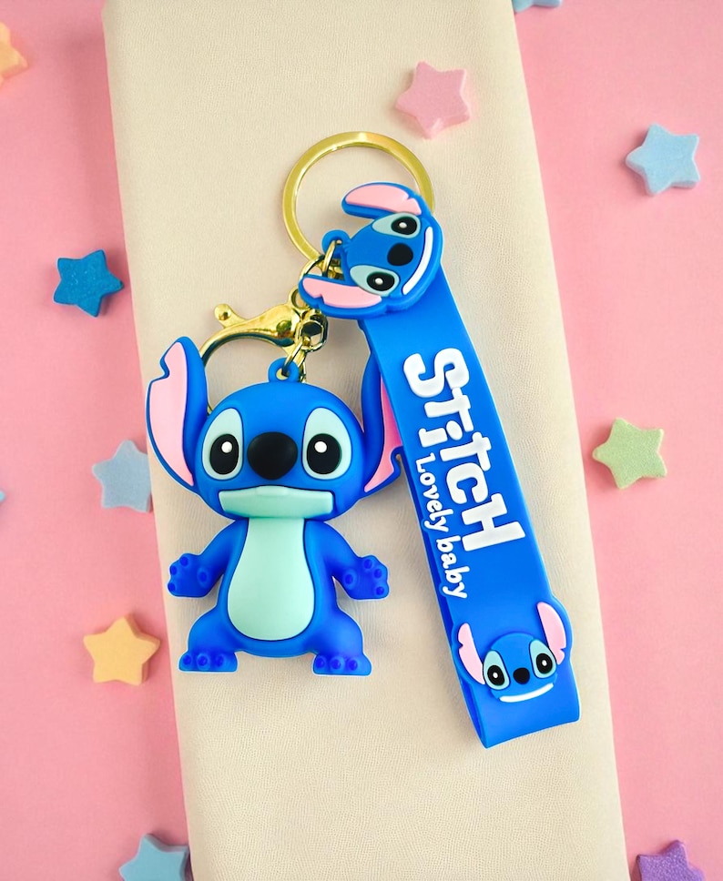 May include: A blue Stitch keychain with a gold-colored keyring and clasp. The keychain features a 3D Stitch figure and a blue strap with the word "Stitch" and "Lovely baby" printed on it. Pink and blue stars are scattered around.