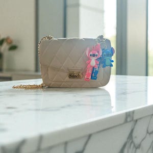 May include: A beige quilted crossbody bag with a gold chain strap. The bag features the word "Stitch" on the front and is adorned with two Stitch character keychains, one pink and one blue.