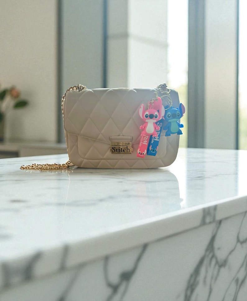 May include: A beige quilted crossbody bag with a gold chain strap. The bag features the word "Stitch" on the front and is adorned with two Stitch character keychains, one pink and one blue.