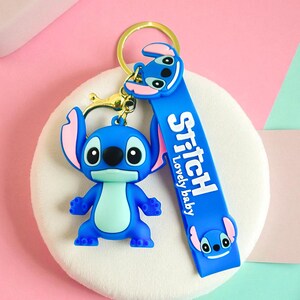 May include: A blue Stitch keychain with a gold-colored keyring and clasp. The keychain features a Stitch figure and a blue strap with the word "Stitch" and "Lovely baby" printed on it.