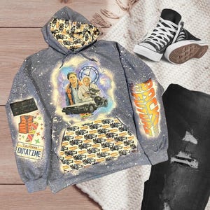 May include: Gray hooded sweatshirt with a bleach-splattered design featuring a scene from the movie Back to the Future. The design includes Marty McFly and Doc Brown standing in front of the DeLorean time machine. The sweatshirt also has a pocket with a repeated pattern of the DeLorean time machine. The text "Back to the Future" is written on the sleeve of the sweatshirt.