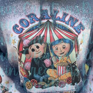 May include: A blue and white bleached hoodie with a graphic of Coraline and the Other Mother from the movie Coraline. The graphic features a circus tent, popcorn, and a black cat. The text "Coraline" is written in a colorful, whimsical font.