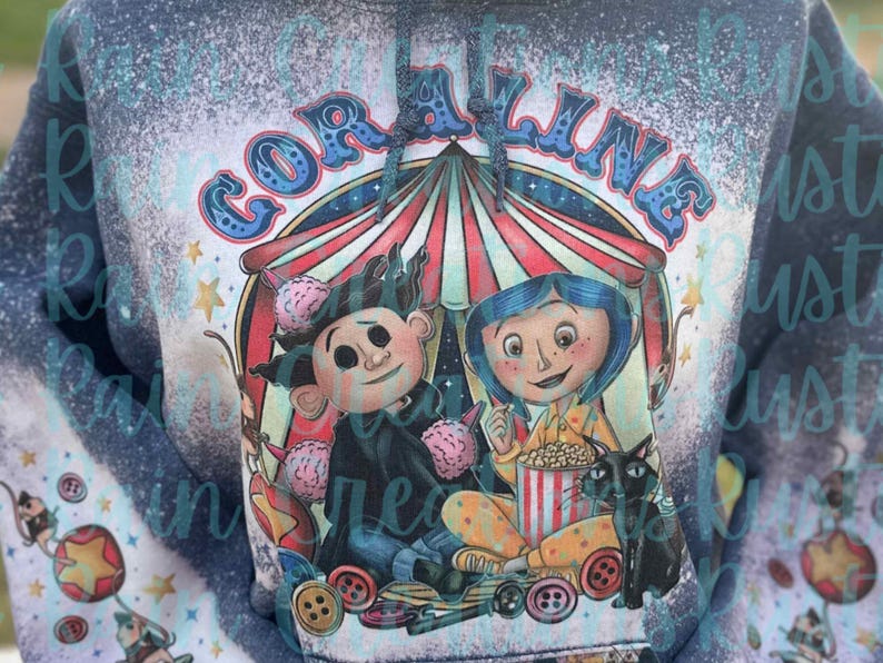 May include: A blue and white bleached hoodie with a graphic of Coraline and the Other Mother from the movie Coraline. The graphic features a circus tent, popcorn, and a black cat. The text "Coraline" is written in a colorful, whimsical font.